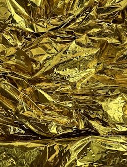 A crumpled sheet of reflective golden material, resembling foil, with numerous creases and folds creating varied reflections and highlights.