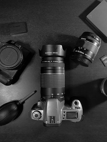 A flat lay arrangement of camera equipment on a dark surface includes a digital camera body, two detachable lenses, a blower brush for cleaning lenses, a hard drive, and two memory cards. The central focus is a camera with an attached zoom lens, complemented by a wide-angle lens to the side. Accessories and storage devices are positioned around the main items, suggesting a theme of photography equipment.