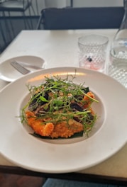 A plate of appetizing food garnished with fresh greens, placed on a white plate. The dish appears to be a type of risotto or creamy puree possibly with pieces of fried or baked ingredients. Surrounding the plate are a glass with a drink, an empty plate, and a fork.