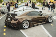 A modified car with a sleek design and prominent rear wing is parked at an outdoor event. Several people in casual clothing are gathered nearby, some appearing to be engaged in conversation. The background includes a white car, a fenced area, and a grassy section with trees.