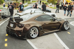 A modified car with a sleek design and prominent rear wing is parked at an outdoor event. Several people in casual clothing are gathered nearby, some appearing to be engaged in conversation. The background includes a white car, a fenced area, and a grassy section with trees.