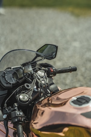 A close-up view of a motorcycle's handlebar area, featuring a digital dashboard with speedometer, a rearview mirror, and several control buttons. The body of the motorcycle has a reflective copper finish, and the background consists of a gravel surface with some blurred greenery.