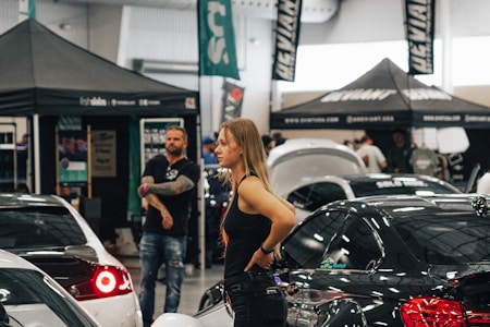 A woman stands among various cars and people at what appears to be an indoor car exhibition or show. Several tents and banners with logos and text are set up in the background. The scene is busy with activity, featuring both males and females looking around and engaging with the cars on display.