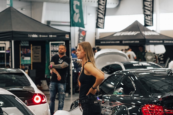 A woman stands among various cars and people at what appears to be an indoor car exhibition or show. Several tents and banners with logos and text are set up in the background. The scene is busy with activity, featuring both males and females looking around and engaging with the cars on display.