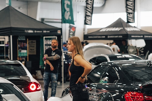 A woman stands among various cars and people at what appears to be an indoor car exhibition or show. Several tents and banners with logos and text are set up in the background. The scene is busy with activity, featuring both males and females looking around and engaging with the cars on display.