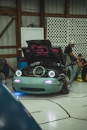 A modified sports car with its hood open, revealing its engine. The car features unique artistic designs on the hood, headlights turned on, and a light teal front bumper. Several people are sitting and engaging in activities around the car inside a garage with wooden structures and lattice panels.