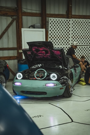 A modified sports car with its hood open, revealing its engine. The car features unique artistic designs on the hood, headlights turned on, and a light teal front bumper. Several people are sitting and engaging in activities around the car inside a garage with wooden structures and lattice panels.
