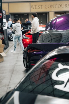 A group of people are gathered around various sports cars in an indoor exhibition hall. The space is well-lit, and several cars with polished exteriors, including a prominent dark purple one, are displayed. The walls feature signage indicating the area is the 'West Concourse.'