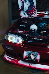 A modified car with its hood open, revealing a stylized engine bay with anime-themed decorations. A poster and a figurine are prominently displayed inside the engine area, both featuring characters with striking red and black color schemes. The front of the car also has anime graphics, including on the license plate.
