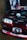 A modified car with its hood open, revealing a stylized engine bay with anime-themed decorations. A poster and a figurine are prominently displayed inside the engine area, both featuring characters with striking red and black color schemes. The front of the car also has anime graphics, including on the license plate.