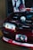 A modified car with its hood open, revealing a stylized engine bay with anime-themed decorations. A poster and a figurine are prominently displayed inside the engine area, both featuring characters with striking red and black color schemes. The front of the car also has anime graphics, including on the license plate.