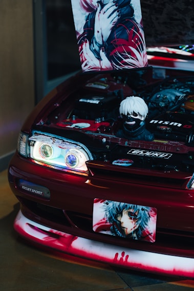 A modified car with its hood open, revealing a stylized engine bay with anime-themed decorations. A poster and a figurine are prominently displayed inside the engine area, both featuring characters with striking red and black color schemes. The front of the car also has anime graphics, including on the license plate.