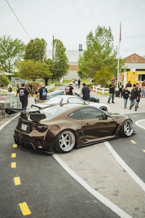 A sleek, modified sports car with an aerodynamic body kit and dark, shiny paint is parked on a winding asphalt track. The scene is lively with several people dressed casually, interacting and admiring the vehicles. The background features a building with a peaked roof and lush green trees, indicating a community or event setting.