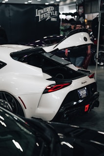 A sleek, white sports car with an open trunk displayed indoors, likely at a car show. A sign reading 'Lowered Lifestyle' is visible at the top. People are gathered around, admiring the vehicle.