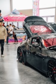A sleek black sports car with custom modifications is displayed indoors. The car's hood is open, revealing intricate detailing, and it features red accents on the wheels. People in casual attire are casually interacting and observing around the car. A vibrant booth with pink and neon colors serves as a backdrop, advertising a brand with banners and signs.