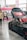A sleek black sports car with custom modifications is displayed indoors. The car's hood is open, revealing intricate detailing, and it features red accents on the wheels. People in casual attire are casually interacting and observing around the car. A vibrant booth with pink and neon colors serves as a backdrop, advertising a brand with banners and signs.