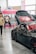A sleek black sports car with custom modifications is displayed indoors. The car's hood is open, revealing intricate detailing, and it features red accents on the wheels. People in casual attire are casually interacting and observing around the car. A vibrant booth with pink and neon colors serves as a backdrop, advertising a brand with banners and signs.