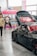 A sleek black sports car with custom modifications is displayed indoors. The car's hood is open, revealing intricate detailing, and it features red accents on the wheels. People in casual attire are casually interacting and observing around the car. A vibrant booth with pink and neon colors serves as a backdrop, advertising a brand with banners and signs.