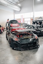 A shiny, modified black sports car is showcased indoors, featuring a custom body kit and red accent lighting in and around the engine compartment. The car's hood is open, revealing the detailed engine work. Other modified cars are visible in the background, along with festival-style tents and banners. The setting appears to be a car show or automotive event inside a spacious venue with high ceilings and industrial lighting.