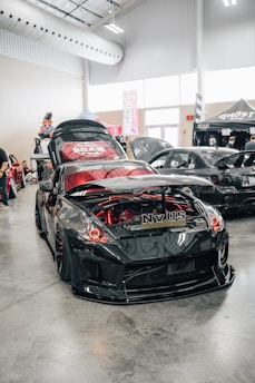 A shiny, modified black sports car is showcased indoors, featuring a custom body kit and red accent lighting in and around the engine compartment. The car's hood is open, revealing the detailed engine work. Other modified cars are visible in the background, along with festival-style tents and banners. The setting appears to be a car show or automotive event inside a spacious venue with high ceilings and industrial lighting.