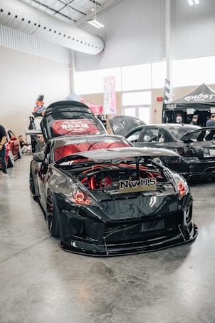 A shiny, modified black sports car is showcased indoors, featuring a custom body kit and red accent lighting in and around the engine compartment. The car's hood is open, revealing the detailed engine work. Other modified cars are visible in the background, along with festival-style tents and banners. The setting appears to be a car show or automotive event inside a spacious venue with high ceilings and industrial lighting.