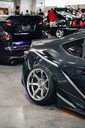 A sleek, modified car is prominently displayed with its polished alloy wheels and distinctive decals. The setting is an indoor car show with several other vehicles and people in the background, including individuals examining the cars and engaging in conversations.