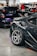 A sleek, modified car is prominently displayed with its polished alloy wheels and distinctive decals. The setting is an indoor car show with several other vehicles and people in the background, including individuals examining the cars and engaging in conversations.