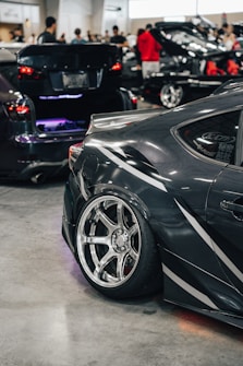 A sleek, modified car is prominently displayed with its polished alloy wheels and distinctive decals. The setting is an indoor car show with several other vehicles and people in the background, including individuals examining the cars and engaging in conversations.