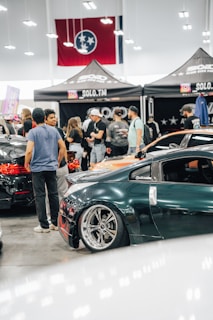 A crowded car show event with several people gathered around sleek, modified cars. The cars are polished with custom paint jobs and shiny rims. A tent with the words 'SOLO.TM' is visible, along with a large flag in the background. The setting is indoors with bright overhead lighting.