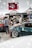 A crowded car show event with several people gathered around sleek, modified cars. The cars are polished with custom paint jobs and shiny rims. A tent with the words 'SOLO.TM' is visible, along with a large flag in the background. The setting is indoors with bright overhead lighting.