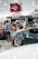 A crowded car show event with several people gathered around sleek, modified cars. The cars are polished with custom paint jobs and shiny rims. A tent with the words 'SOLO.TM' is visible, along with a large flag in the background. The setting is indoors with bright overhead lighting.