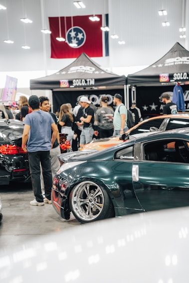 A crowded car show event with several people gathered around sleek, modified cars. The cars are polished with custom paint jobs and shiny rims. A tent with the words 'SOLO.TM' is visible, along with a large flag in the background. The setting is indoors with bright overhead lighting.