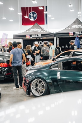 A crowded car show event with several people gathered around sleek, modified cars. The cars are polished with custom paint jobs and shiny rims. A tent with the words 'SOLO.TM' is visible, along with a large flag in the background. The setting is indoors with bright overhead lighting.