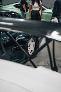 A close-up view of a car's wheel with distinctive rims is partially obscured by what appears to be a wing or spoiler. In the background, several people are standing near another vehicle, which is green. The scene appears to be indoors, possibly at a car show or exhibition.
