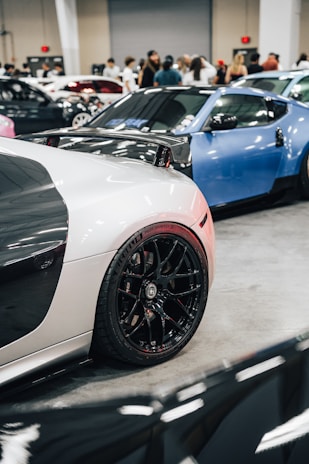 A car exhibition featuring high-end sports cars. The foreground focuses on a black and white sports car with shiny black rims, while a blue car is parked beside it. People are gathered in the background, observing the vehicles within an indoor setting with bright lighting.