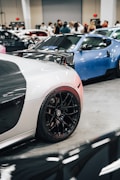 A car exhibition featuring high-end sports cars. The foreground focuses on a black and white sports car with shiny black rims, while a blue car is parked beside it. People are gathered in the background, observing the vehicles within an indoor setting with bright lighting.