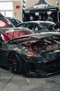 A sleek, modified black sports car is on display indoors, its hood open to showcase the customized engine with red components. Nearby, a tent with the name 'Ghost Gang' is set up, and in the background, there are a few people and another car visible.