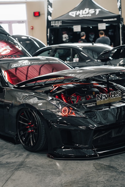 A sleek, modified black sports car is on display indoors, its hood open to showcase the customized engine with red components. Nearby, a tent with the name 'Ghost Gang' is set up, and in the background, there are a few people and another car visible.