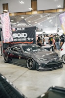 A custom car show with a sleek, dark gray sports car prominently displayed. The car has black tinted windows and low-profile tires with shiny rims. Surrounding the car are banners and canopies with logos and branding, indicating a promotional setup. A crowd of people in casual attire gathers around, engaging with the exhibits.