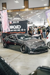 A custom car show with a sleek, dark gray sports car prominently displayed. The car has black tinted windows and low-profile tires with shiny rims. Surrounding the car are banners and canopies with logos and branding, indicating a promotional setup. A crowd of people in casual attire gathers around, engaging with the exhibits.