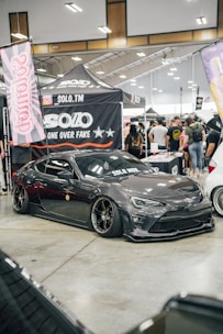 A custom car show with a sleek, dark gray sports car prominently displayed. The car has black tinted windows and low-profile tires with shiny rims. Surrounding the car are banners and canopies with logos and branding, indicating a promotional setup. A crowd of people in casual attire gathers around, engaging with the exhibits.