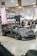 A custom car show with a sleek, dark gray sports car prominently displayed. The car has black tinted windows and low-profile tires with shiny rims. Surrounding the car are banners and canopies with logos and branding, indicating a promotional setup. A crowd of people in casual attire gathers around, engaging with the exhibits.