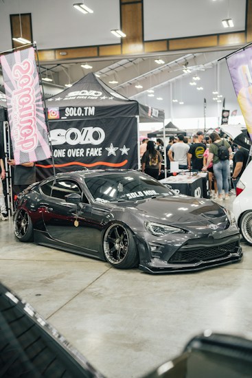 A custom car show with a sleek, dark gray sports car prominently displayed. The car has black tinted windows and low-profile tires with shiny rims. Surrounding the car are banners and canopies with logos and branding, indicating a promotional setup. A crowd of people in casual attire gathers around, engaging with the exhibits.
