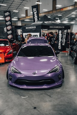 A purple Toyota sports car is prominently displayed indoors at a car show. Surrounding the car are several banners and a canopy with branding for various automotive companies. People are gathered in the background, engaging with displays and each other. The environment is well-lit with overhead lights visible on the ceiling.