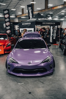 A purple Toyota sports car is prominently displayed indoors at a car show. Surrounding the car are several banners and a canopy with branding for various automotive companies. People are gathered in the background, engaging with displays and each other. The environment is well-lit with overhead lights visible on the ceiling.