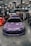 A purple Toyota sports car is prominently displayed indoors at a car show. Surrounding the car are several banners and a canopy with branding for various automotive companies. People are gathered in the background, engaging with displays and each other. The environment is well-lit with overhead lights visible on the ceiling.