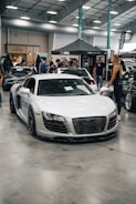 A sleek white sports car is displayed indoors at an automotive event. Several people are standing nearby, interacting and observing the cars. A tent with branding is in the background, along with other luxury vehicles parked in a spacious area with a high ceiling.