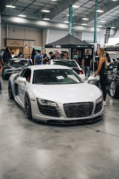 A sleek white sports car is displayed indoors at an automotive event. Several people are standing nearby, interacting and observing the cars. A tent with branding is in the background, along with other luxury vehicles parked in a spacious area with a high ceiling.