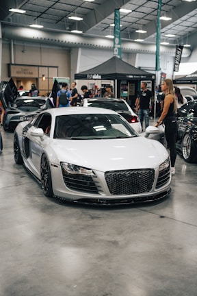 A sleek white sports car is displayed indoors at an automotive event. Several people are standing nearby, interacting and observing the cars. A tent with branding is in the background, along with other luxury vehicles parked in a spacious area with a high ceiling.