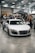 A sleek white sports car is displayed indoors at an automotive event. Several people are standing nearby, interacting and observing the cars. A tent with branding is in the background, along with other luxury vehicles parked in a spacious area with a high ceiling.